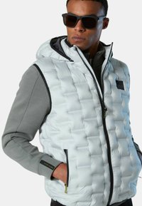 Gepolsterte, hellgraue Weste mit Kapuze, ausgestattet mit einem Reißverschluss vorne, schwarzem Besatz, Seitentaschen und einem strukturierten, gesteppte Design.