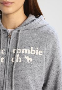 Šedá mikina s kapucí z texturované látky, s vyšívaným bílým textem a grafikou losa. Obsahuje zip a stahovací šňůrku na kapuci.