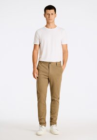Pantaloni beige su misura con una texture liscia, chiusura frontale con bottone, tasche laterali, indossati con una t-shirt bianca semplice e scarpe da ginnastica bianche.