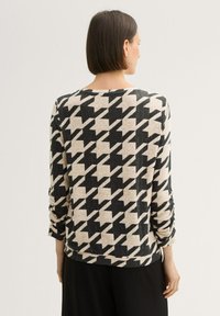 Dugmač s dugim rukavima s uzorkom houndstooth u crnoj i bež boji. Mekana, teksturirana tkanina s okruglim izrezom i blago opuštenim krojem.
