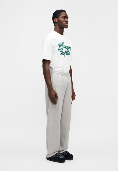 Λευκό μπλουζάκι με το πράσινο κεντητό λογότυπο "Billionaire Boys Club", συνδυασμένο με γκρι ανοιχτό χρώμα φόρμες και μαύρα παντόφλες με σχέδια.