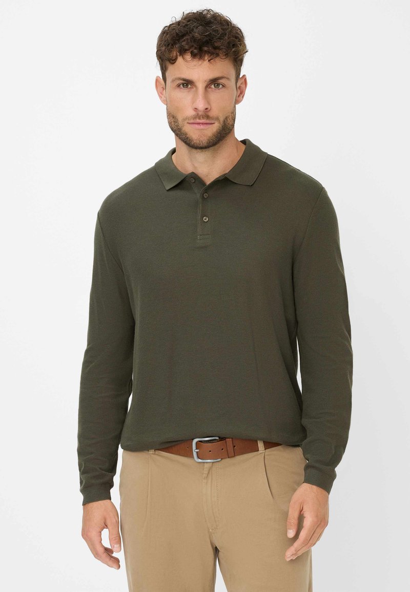 Dunkelgrünes langärmliges Poloshirt mit Kragen, einer Knopfleiste mit drei Knöpfen und gerippten Manschetten, kombiniert mit hellbeigen Hosen und einem braunen Gürtel.