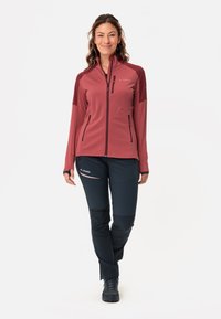Giacca softshell bordeaux con zip frontale e dettagli neri; abbinata a pantaloni neri aderenti con dettagli rosa e calzature nere robuste.