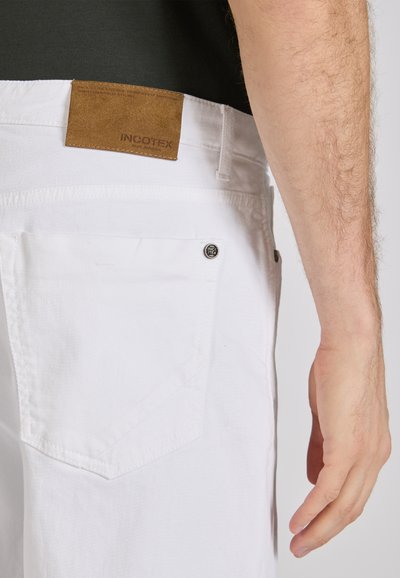 Pantalones de algodón blancos con una etiqueta de marca de cuero marrón, con un bolsillo trasero y un botón de logo metálico. Textura suave y corte ajustado.