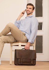 Brun læder dokumentmappe med spænder, rektangulær form, glat overflade; mand i lyseblå skjorte sidder på en træstol.