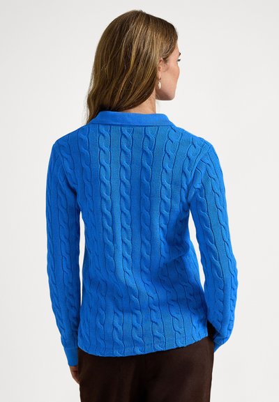 Moteris dėvi ryškiai mėlyną megztinį su pynimais ir apykakle, stovinti nugara į žiūrovą priešais paprastą šviesų foną.