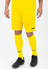 Gelbe Sportshorts und knielange Socken, aus synthetischem Material. Schwarzes JAKO-Logo auf Shorts und Socken. Schwarze Fußballschuhe mit strukturiertem Design.