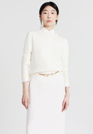 Donna che indossa un maglione bianco sopra una camicia bianca con colletto, una cintura dorata e una gonna bianca, in piedi di fronte a uno sfondo chiaro e semplice.