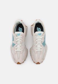 Pár béžových a světle modrých tenisek New Balance s bílými tkaničkami, pohled shora na bílém pozadí.