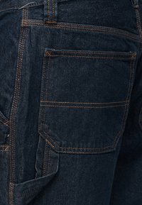 Κοντινό πλάνο σκούρου μπλε τζιν με πίσω τσέπη που έχει πορτοκαλί ραφές και πλαϊνό λουράκι χρησιμότητας.