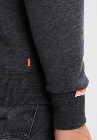 Dark grey sweatshirt with ribbed cuffs, featuring a small orange tag with "Superdry" branding on the sleeve. Soft, textured fabric.