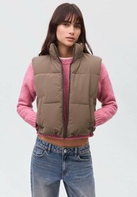 Gilet imbottito marrone con colletto alto, chiusura con zip e tasche laterali. Indossato sopra un maglione rosa, abbinato a jeans blu. Texture liscia.