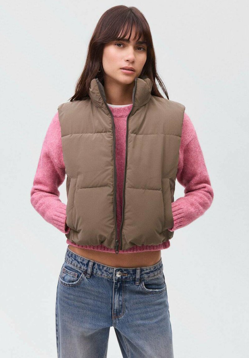 Gilet imbottito marrone con colletto alto, chiusura con zip e tasche laterali. Indossato sopra un maglione rosa, abbinato a jeans blu. Texture liscia.