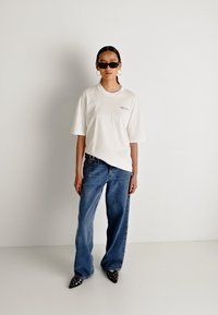 Camiseta blanca de algodón con corte holgado, combinada con vaqueros de pierna ancha en azul deslavado. Los accesorios incluyen gafas de sol negras y zapatos de punta con embellishments.