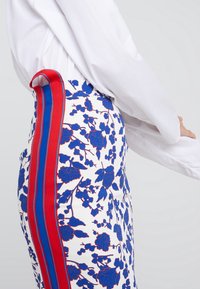 Květinová sukně s modrými a červenými akcenty na bílém pozadí. Boční panel má výrazný červený pruh vedle květinového vzoru.