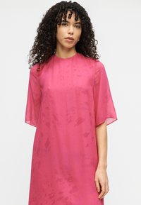 Ροζ διάφανο φόρεμα με κοντά μανίκια, που διαθέτει άνετη εφαρμογή και διακριτικά υφασμάτινα σχέδια. Αέρινο ύφασμα που πέφτει κομψά.