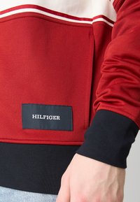 Felpa rossa con una texture uniforme, caratterizzata da un polsino nero e un design a strisce bianche e rosse. Un piccolo etichetta nera mostra la scritta "HILFIGER."