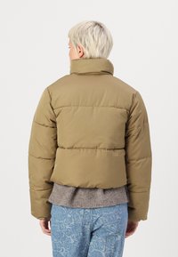 Vihreä puffatakki korkealla kauluksella, laatikkomainen siluetti ja vaHorizontal stitching. Teksturoitu harmaa neulepaidasta näkyy helmassa, yhdistettynä sinisiin kuviollisiin housuihin.