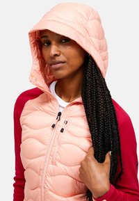 Chaqueta plumón de color rosa claro con textura acolchada, presenta una capucha y cierre de cremallera. Las mangas rojas aportan contraste al diseño.