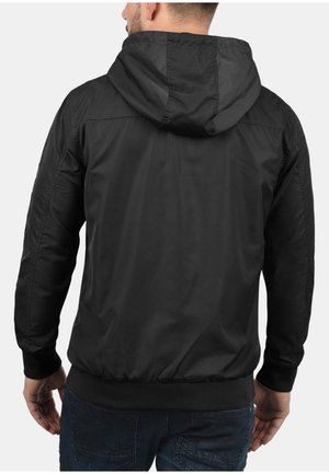 Man wearing a black hooded jacket with elastic cuffs and waistband, shown from the back against a plain background.