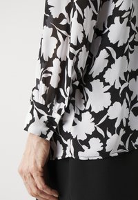 Svart och vit blommig blus med genomskinlig textur, långa ärmar och knäppte manschetter. Mönstret har stora, abstrakta blomformer.