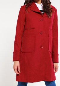 Abrigo de mezcla de lana rojo con cuello de muesca, cierre de botones, dos bolsillos frontales y una silueta entallada. Mangas largas con detalles en los puños.