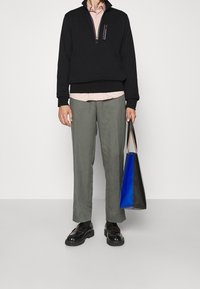 Sweatshirt zippé noir avec une poche accentuée de violet, chemise rose claire en dessous, pantalon vert olive et un sac fourre-tout bleu-noir.