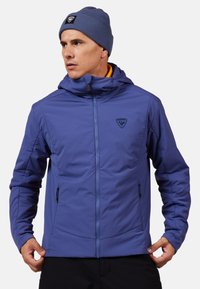 Lila gepolsterte Jacke mit einem Reißverschluss vorne, einer Kapuze und zwei seitlichen Taschen. Sie hat ein kleines Logo auf der Brust und eine glatte, strukturierte Oberfläche.