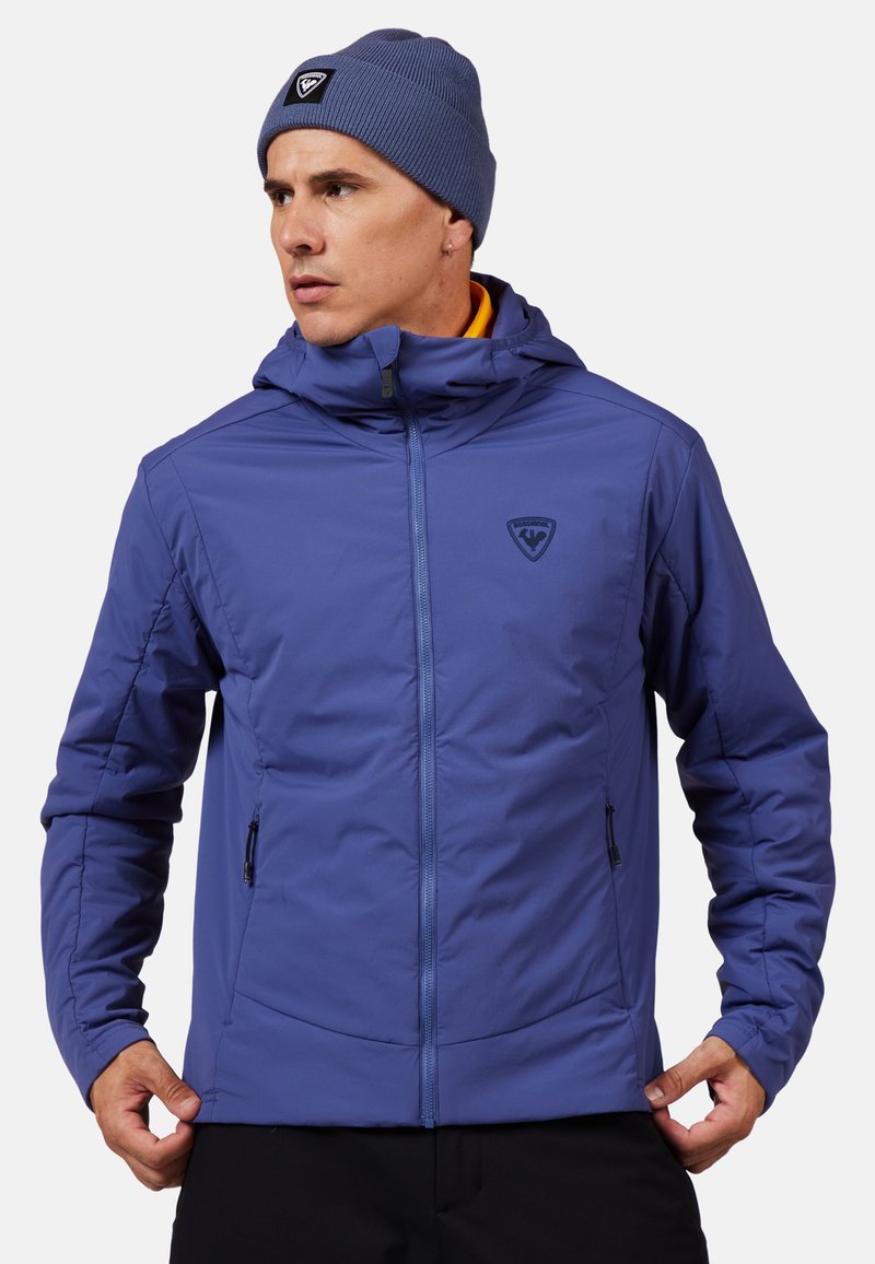 Lila gepolsterte Jacke mit einem Reißverschluss vorne, einer Kapuze und zwei seitlichen Taschen. Sie hat ein kleines Logo auf der Brust und eine glatte, strukturierte Oberfläche.