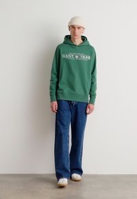 Felpa verde con stampa del logo bianco, abbinata a jeans a gamba larga blu e sneakers bianche. Modello con un berretto crema, in piedi davanti a uno sfondo neutro.