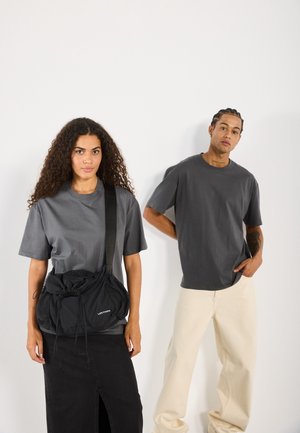 Jeune femme aux cheveux bouclés portant un t-shirt gris, une jupe noire et un sac bandoulière noir ; jeune homme aux cheveux tressés portant un t-shirt gris et un pantalon beige.