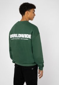 Groene sweatshirt met witte blokletters "WORLDWIDE" en extra tekst op de achterkant. Heeft een relaxte pasvorm en ribgebreide manchetten.