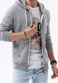 Sweat à capuche gris à fermeture éclair avec poignets et ourlet côtelés, doté de poches avant, porté par-dessus un t-shirt blanc à imprimé graphique. Jeans en denim décontractés visibles.