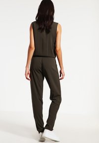 Olivgrüner ärmelloser Jumpsuit mit lockerer Passform, schmal zulaufenden Beinen und glatter Textur. Getragen mit weißen Sneakers, die ein schlichtes Design aufweisen.