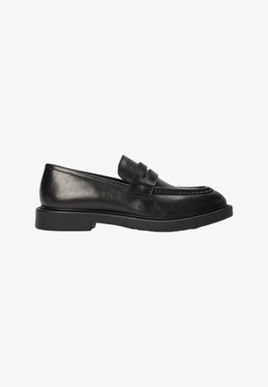 Mocassin en cuir noir avec un bout arrondi et une semelle en caoutchouc basse, présentant une sangle décorative sur le dessus et des détails de couture subtils.