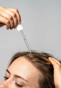 Klar dropper med en hvid hætte, der dispenserer væske på brunt hår ved hårbunden. Lyst gråt baggrund, teksturerede hårstrå synlige.