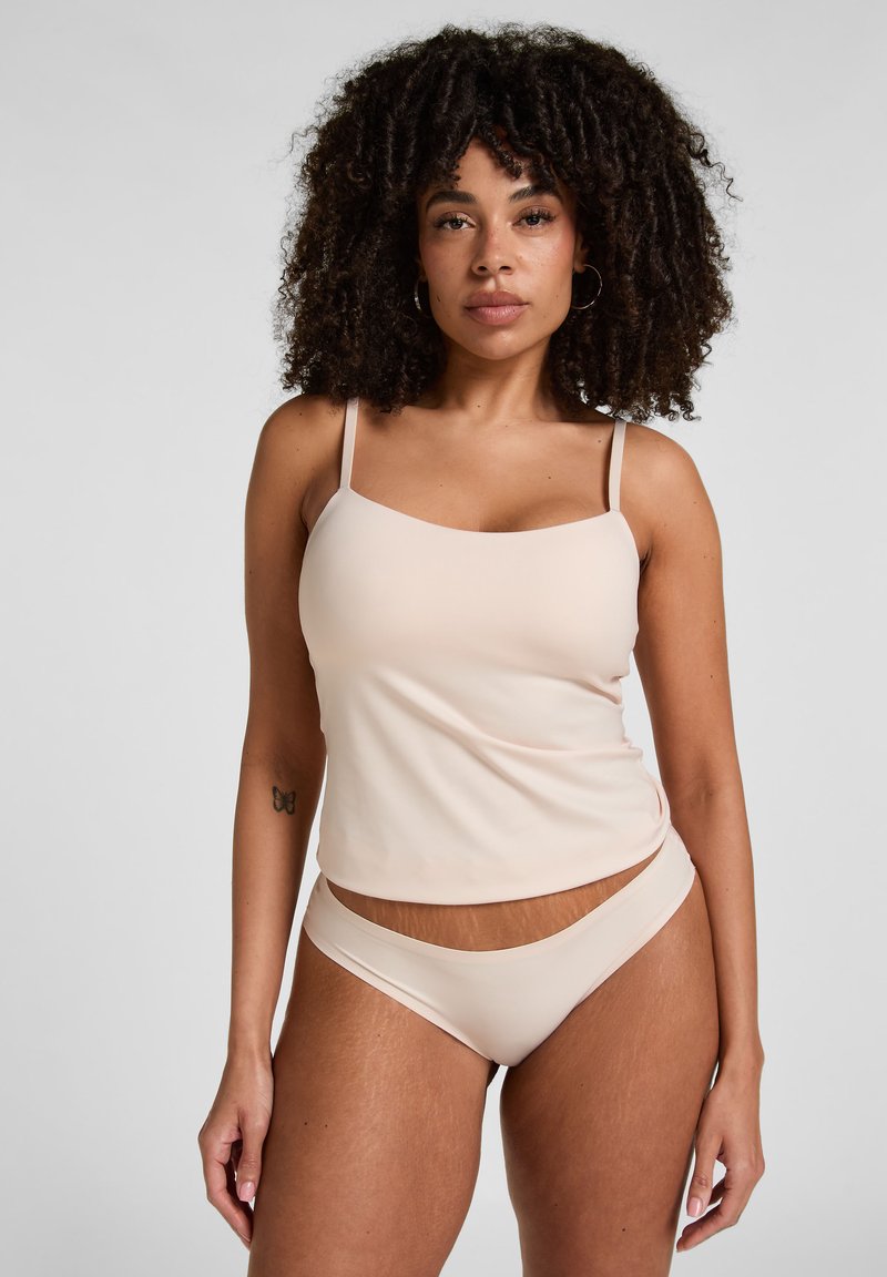 Femme aux cheveux bouclés portant un caraco beige et des sous-vêtements assortis, debout devant un fond gris uni, faisant face à l'avant.