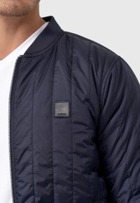 Mørkeblå quilted bomberjakke med ribbet krave og lynlås. Har et lille sort mærke på venstre bryst. Glat syntetisk tekstur.