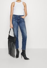 Mujer vestida con una blusa sin mangas blanca, jeans azules con adornos laterales, botas de tacón alto negras y puntiagudas hasta el tobillo, sosteniendo un bolso de flecos negro.