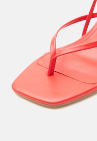 Κοντινή λήψη σανδαλιού σε κοραλλί χρώμα με τετράγωνη μύτη και λεπτά λουράκια σε απλό φόντο.