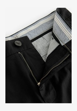 Un pantalon noir en coton dispose d'une fermeture éclair et d'un bouton. La taille est dotée d'une doublure à motifs avec des rayures et des accents texturés.
