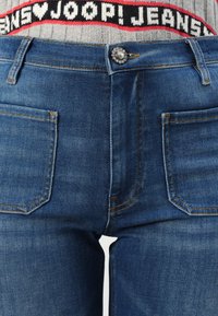 Blaue Denim-Jeans mit einem vorderen Knopfverschluss, zwei Taschen vorne und kontrastierendem orangefarbenem Nähten. Der Bund besitzt einen markierten Taillengummi.
