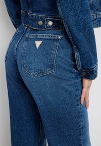Jeans en denim bleu moyen, avec un patch logo triangulaire sur la poche arrière, des coutures standard et une coupe classique.