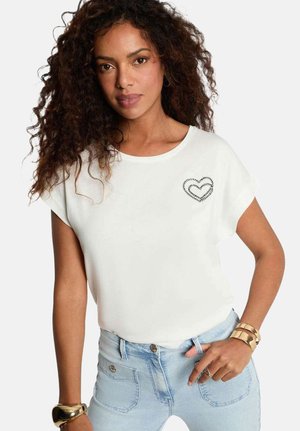 Femme aux cheveux bouclés portant une chemise blanche avec un cœur cousu sur la poitrine, un jean bleu clair et des bracelets dorés au poignet.