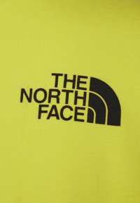 Tessuto giallo brillante con un logo nero che recita "THE NORTH FACE", caratterizzato da una forma di mezza luna stilizzata e una grafica in caratteri bold.