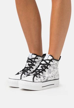 Witte hoge sneakers met zwarte abstracte lijnpatronen en zwarte veters, gedragen door een persoon met blote benen tegen een effen achtergrond.