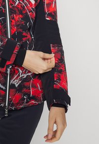 Röd och svart mönstrad jacka med dragkedja fram, texturerade ärmadetaljer och kontrasterande svarta manschettdetaljer.