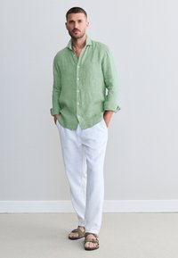Chemise en lin vert clair avec col, pantalon en lin blanc, et sandales vert olive. Coupe décontractée, manches longues retroussées aux poignets.