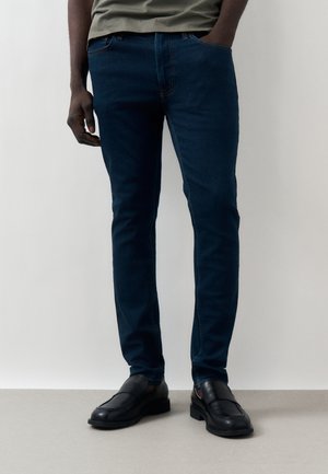 Άνδρας που φοράει σκουρό μπλε παντελόνι τζην στενής εφαρμογής, πράσινο πουκάμισο, μαύρα δερμάτινα μοκασίνια, στέκεται με το ένα χέρι στην τσέπη μπροστά σε μονόχρωμο φόντο.