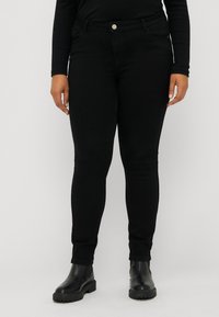 Jean skinny noir avec une texture lisse, doté de poches avant, d'une fermeture à bouton, et d'un design ajusté. Porté avec des bottes noires épaisses.